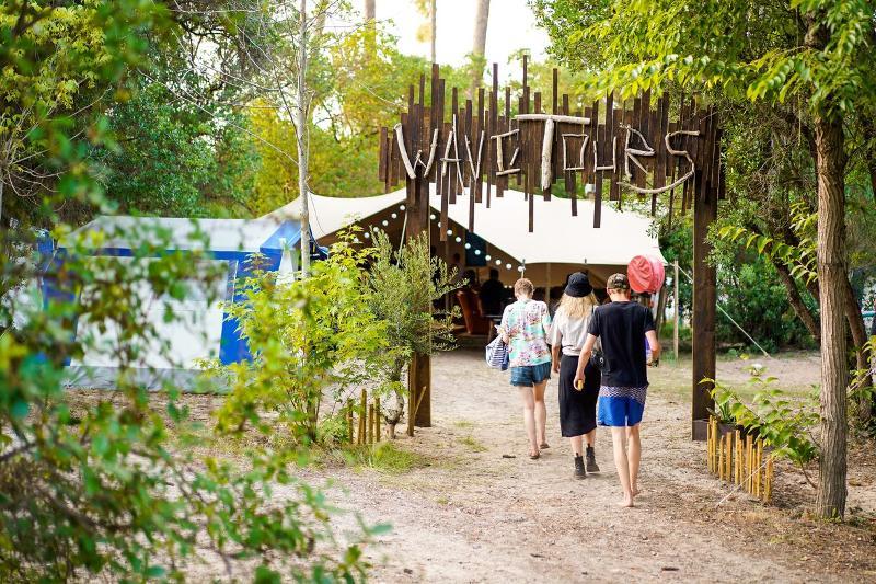  três jovens a caminhar por um trilho em direção a uma tenda, passando por uma entrada rústica feita de madeira com a palavra 
