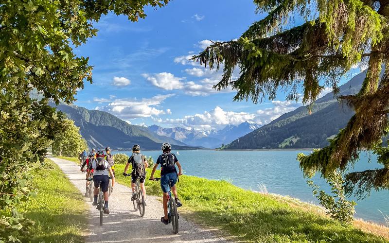 Teilnehmer eines MTB-Camps fahren hintereinander an einem blauen See in den Alpen