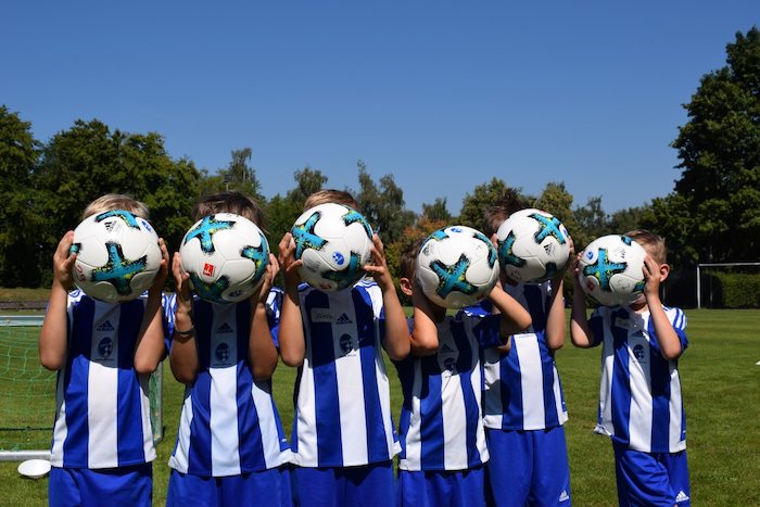 Im Englischcamp Pfingstferien stehen die Kinder auf einem Fußballplatz und halten sich einen Ball vor den Kopf