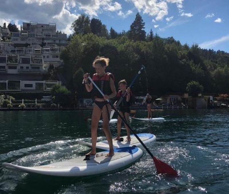 Zwei Teilnehmerinnen üben das SUP Board paddeln auf einem See in ihren Lernferien