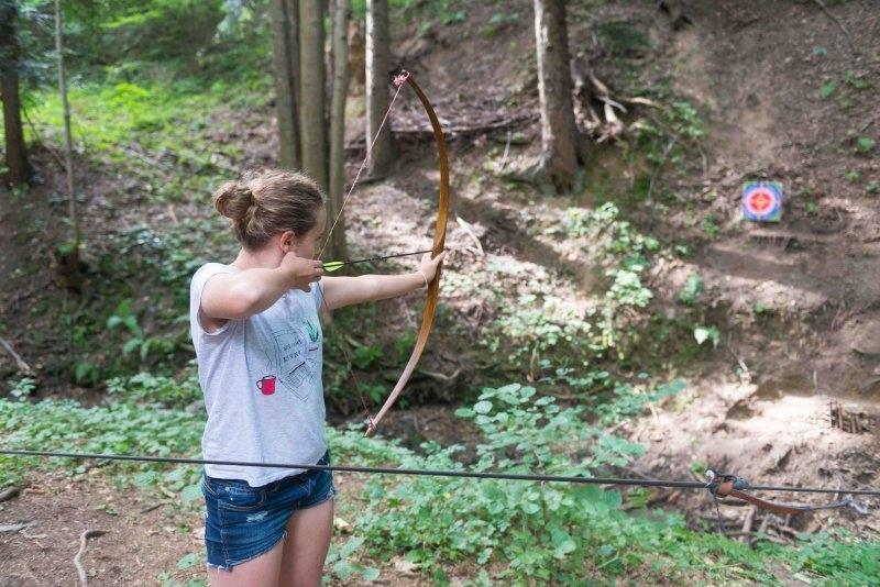 Mädchen steht während ihrer Kinderbetreuung Sommerferien im Wald und schießt einen Bogen auf eine Zielscheibe