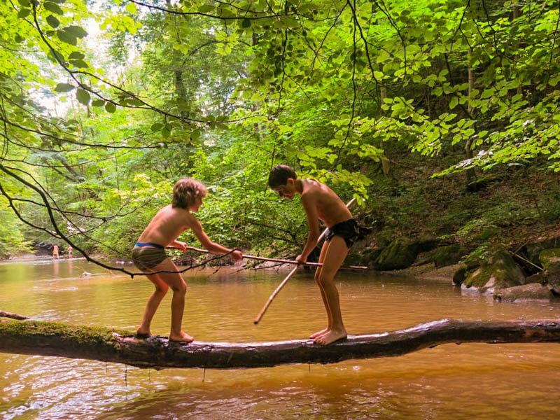 Zwei Jungs beim Schwertkampf mit Holzstöcken über einen Fluss im Sommercamp Oberösterreich