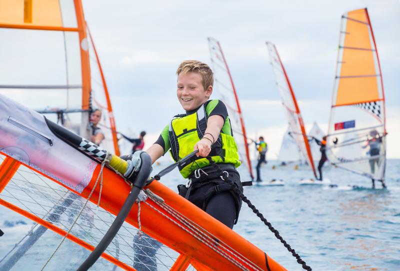 Ein blonder Junge hat viel Spaß beim Windsurfen im Wassersportcamp