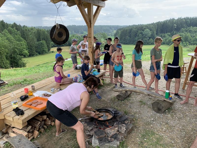  Num cenário de campo na Suécia, um grupo de jovens forma uma fila para receber comida enquanto uma mulher prepara hambúrgueres numa grelha ao ar livre, protegida por um telhado de madeira. Ao fundo, estendem-se campos verdes e uma floresta, criando um ambiente acolhedor e natural perfeito para um campo de férias suécia 
