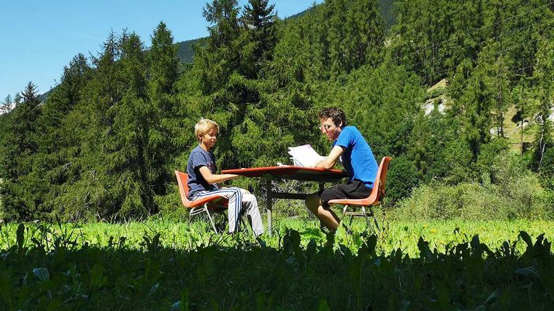 Ein Junge und eine Lehrkraft sitzen an einem Tisch umgeben von Bäumen und lernen zusammen