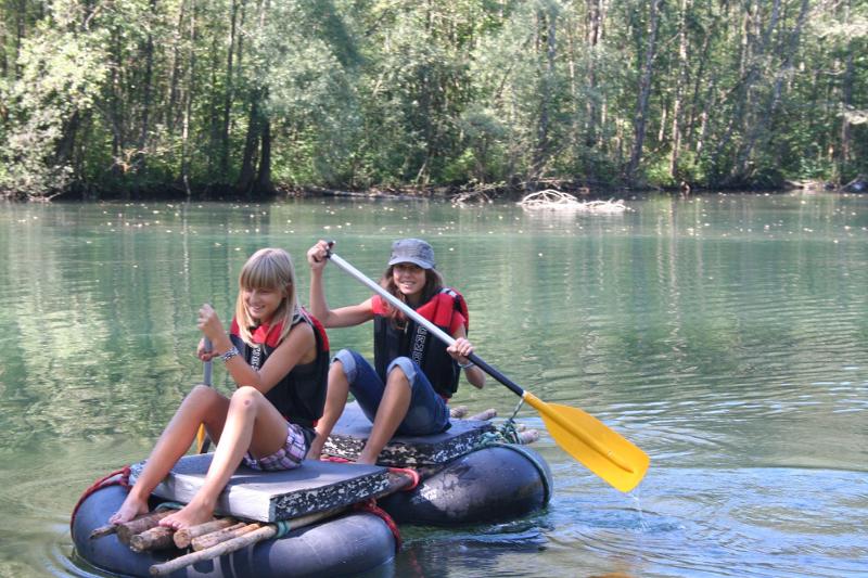 Zwei Mädchen sitzen auf einem selbstgebauten Floß und paddeln im Actioncamp durch den Fluss