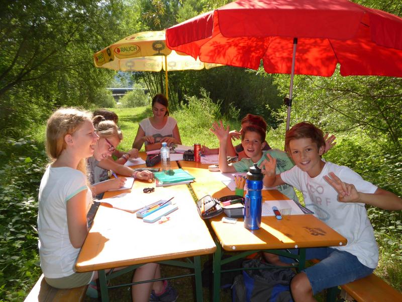 Teilnehmer eines Kindersprachkurses sitzen im freien an einem Tisch und lernen Sprachen
