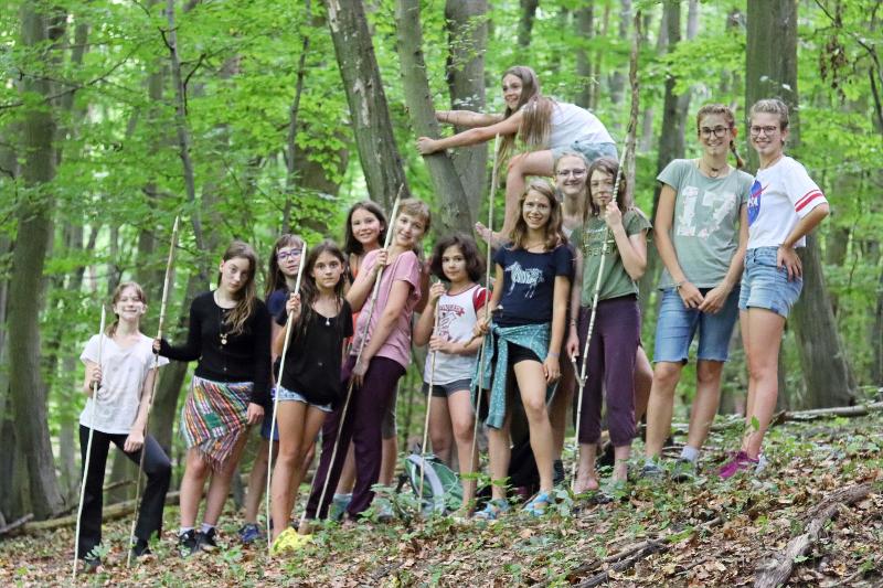13 Mädchen stehen mit Stöckern im Wald und lächeln in die Kamera