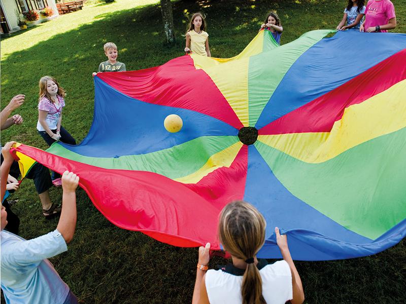 Die Kinder eines Feriencamp Linz stehen im Kreis und spielen mit Bällen und einem buntem Tuch